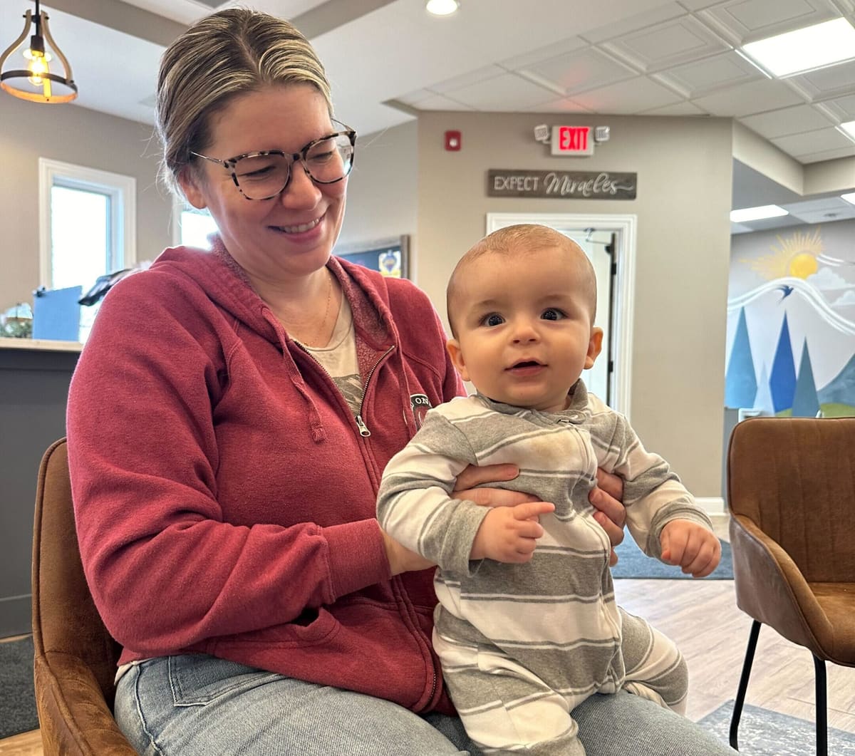 Mother holding an infant in an office waiting area with an 'Expect Miracles' wall sign behind them