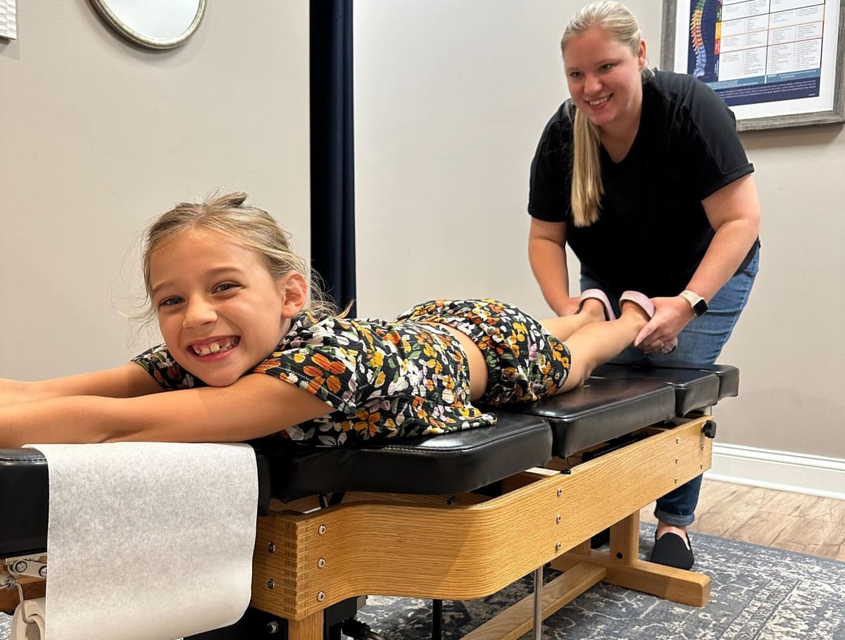 A woman gently holds a smiling girl's ankles as the girl lies face down on a treatment table.
